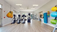 This fitness center in Memphis, TN offers a thoughtfully designed, single-floor workout facility that balances functionality with an energizing atmosphere. The space is anchored by a striking white brick accent wall adorned with a bold Muhammad Ali quote — "Float Like a Butterfly, Sting Like a Bee" — alongside a pair of decorative boxing gloves, setting an motivational tone throughout.

The main workout area features a generous row of Matrix treadmills and elliptical cross-trainers positioned to take advantage of large floor-to-ceiling windows and glass garage-style doors, which flood the space with abundant natural light and provide views of the surrounding outdoor landscape. Light wood-grain flooring runs throughout, contributing to a clean and modern aesthetic.

A dedicated functional training zone includes a multi-use cable and strength machine, resistance bands, stability balls in multiple sizes, a dip/pull-up station, free weights, and a flat bench — catering to a wide range of workout styles. A ceiling fan provides additional airflow across the open floor plan, complemented by overhead fluorescent lighting.

Along the perimeter, organized storage for accessories and signage keeps the space tidy and user-friendly. A separate, partially enclosed area appears to serve as a resource or stretch station, adding further versatility. The overall ambiance is bright, clean, and welcoming, making it a comprehensive facility for cardio, strength, and functional fitness training.