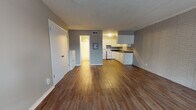 This single-floor unit at 2424 NW 39th St in Oklahoma City offers a practical, open layout with notable architectural character. Wood-style flooring flows consistently throughout the main living areas, complementing the neutral gray walls and textured ceilings.

The living area is a generous open space anchored by a distinctive painted brick accent wall that adds texture and visual interest. A window along the exterior wall allows natural light to filter in, and a wall-mounted AC unit provides climate control. The front entry door opens directly into this room, making the layout straightforward and accessible.

The kitchen connects seamlessly to the living area and features white cabinetry with ample upper and lower storage, dark countertops, a stainless steel double sink, a white electric range, and a full-size refrigerator. A tile backsplash and the continuing brick wall add dimension to the cooking space.

A hallway connects the main living area to the bathroom and additional rooms. The bathroom is well-appointed with a white vanity featuring a speckled quartz-style countertop, an integrated sink, a large mirror, and a combined tub and shower with white tile surround. Wood-style flooring carries into the bathroom, maintaining a cohesive look throughout the unit.