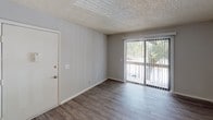 This single-floor residential unit offers a practical and well-organized layout designed for comfortable everyday living. The galley-style kitchen is efficiently arranged with white cabinetry lining both sides, ample counter space with light-toned laminate surfaces, an electric coil range, and a sink — all set atop durable wood-look flooring that adds warmth to the space. The kitchen connects seamlessly to the living area, which features the same wood-look flooring, neutral gray walls, and a textured ceiling. A sliding glass door with vertical blinds fills the living room with natural light and provides direct access to an outdoor patio, offering a pleasant view of surrounding greenery. Adjacent to the living area, the bedroom is finished with plush carpet, a window with blinds that allows natural light in, and a textured ceiling, creating a quiet and comfortable retreat. The bathroom is appointed with a full tub-shower combo, a single vanity with under-sink storage, a mirror, and a light bar, all set against warm gray walls and wood-look flooring. Throughout the unit, a consistent neutral color palette and clean finishes create a cohesive and move-in-ready environment.