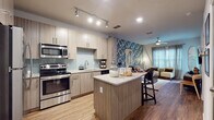 Located at 2855 Gulf to Bay Blvd in Clearwater, Florida, this thoughtfully designed single-floor residence blends modern finishes with a vibrant coastal aesthetic. The open-concept kitchen is a standout feature, equipped with stainless steel appliances including a range, microwave, dishwasher, and refrigerator. Light wood-grain cabinetry pairs elegantly with a soft blue subway tile backsplash and granite-topped island, which doubles as a breakfast bar with seating. Track lighting overhead adds a contemporary touch, while wood-look flooring flows seamlessly throughout the space. The living area is defined by a bold tropical-leaf accent wall that gives the room a striking, resort-inspired character. A ceiling fan, natural light from large windows with sheer curtains, and curated coastal artwork — including surf-themed pieces — complete the relaxed yet stylish ambiance. The bedroom offers a tranquil retreat with warm, muted wall tones, a ceiling fan, natural light, and wood-look flooring. The full bathroom features a bathtub and shower combination, a long vanity with ample storage, and clean, neutral finishes that complement the overall design palette of the residence.
