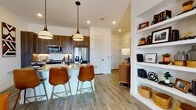 This thoughtfully designed single-floor residence offers 1 bedroom and 1 bathroom, combining modern aesthetics with warm, eclectic details. The open-concept kitchen is a standout, featuring rich walnut-toned cabinetry, stainless steel appliances, a large center island with a light quartz countertop, and stylish pendant lighting overhead. Leather-upholstered barstools with gold-toned legs anchor the island, making it an ideal gathering spot. Light wood-look flooring runs throughout the main living areas, contributing to a bright, cohesive feel. Built-in open shelving provides both storage and a display area, adding character to the living space. The living room is warm and inviting, centered around a tufted charcoal sofa and complemented by a gallery wall arrangement and lush greenery. The bedroom is a serene retreat, distinguished by a dramatic reclaimed wood accent wall that adds rustic texture. Large windows allow generous natural light to fill the room while offering outdoor views, and plush carpet underfoot enhances comfort. The bathroom features wood-look tile flooring, granite countertops with a walnut vanity, a glass-enclosed shower, and convenient access to a walk-in closet with ample rod storage. Throughout the residence, the design balances contemporary finishes with warm, natural materials for a cohesive and inviting ambiance.