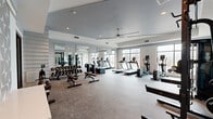 This modern fitness center offers a comprehensive workout environment thoughtfully designed across a single open floor. The space is bright and airy, benefiting from large, floor-to-ceiling windows along multiple walls that flood the interior with natural light and provide views of the surrounding exterior. A tray ceiling with a ceiling fan enhances the sense of vertical space and ensures comfortable airflow throughout.

The layout is organized to accommodate a wide range of fitness activities. The cardio zone features multiple treadmills, elliptical trainers, and a rowing machine, all positioned to take advantage of the natural light from the expansive windows. The strength training area is well-appointed with a full dumbbell rack, adjustable benches, cable machines, and squat racks complete with weight plates — catering to users of all fitness levels.

Full-length mirrors line an entire wall, visually expanding the space while serving a functional role for form monitoring during exercise. The flooring combines carpet and rubber-style surfaces appropriate for both machine use and free weight training. Recessed lighting complements the natural light, ensuring the space is well-illuminated throughout the day. The overall ambiance is clean, contemporary, and professional, reflecting a high-quality amenity facility.