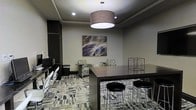 This single-floor space functions as a versatile business lounge or clubroom, thoughtfully designed to accommodate both focused work and casual gathering. The room features a tray ceiling with recessed lighting complemented by a prominent large drum pendant light that anchors the space and casts a warm, inviting glow throughout. The walls are finished in neutral gray tones, providing a sophisticated backdrop enhanced by a bold abstract canvas artwork that serves as a visual focal point. The flooring consists of patterned multi-tone carpet tiles, adding texture and a contemporary feel underfoot. A dark espresso-toned high-top communal table with sleek metal bar stools occupies the central area, ideal for collaborative work or informal meetings. Along one wall, a dedicated workstation area features built-in shelving and individual desks with modern chairs, offering a practical setup for computer use or individual tasks. Complementing the workspace, a pair of patterned accent chairs with small side tables provides a comfortable seating nook for relaxed conversation or reading. Decorative wire cube sculptures add an artistic touch, while the overall design maintains a clean, professional ambiance well-suited for a shared amenity space.