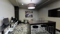 This single-floor space functions as a versatile business lounge or clubroom, thoughtfully designed to accommodate both focused work and casual gathering. The room features a tray ceiling with recessed lighting complemented by a prominent large drum pendant light that anchors the space and casts a warm, inviting glow throughout. The walls are finished in neutral gray tones, providing a sophisticated backdrop enhanced by a bold abstract canvas artwork that serves as a visual focal point. The flooring consists of patterned multi-tone carpet tiles, adding texture and a contemporary feel underfoot. A dark espresso-toned high-top communal table with sleek metal bar stools occupies the central area, ideal for collaborative work or informal meetings. Along one wall, a dedicated workstation area features built-in shelving and individual desks with modern chairs, offering a practical setup for computer use or individual tasks. Complementing the workspace, a pair of patterned accent chairs with small side tables provides a comfortable seating nook for relaxed conversation or reading. Decorative wire cube sculptures add an artistic touch, while the overall design maintains a clean, professional ambiance well-suited for a shared amenity space.