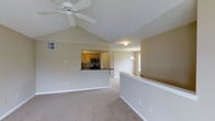 This single-floor home is defined by its open-concept design, where the living area and kitchen flow seamlessly together to create a spacious, cohesive environment. The living area benefits from vaulted ceilings that rise to a prominent peak, amplifying the sense of openness and airiness throughout the space. A ceiling fan is centrally mounted at the apex of the vaulted ceiling, providing both function and a finishing architectural detail.

Neutral-toned carpet covers the living area floors, contributing to a warm and inviting atmosphere. The walls are finished in a soft, light neutral tone that complements the bright, airy feel of the space.

The kitchen is accessible via a half-wall pass-through, which maintains an open visual connection between the two areas while defining each space. The kitchen features warm honey-toned wood cabinetry paired with dark countertops and stainless steel appliances, including a microwave and range. Adjacent to the kitchen, a hallway leads toward additional areas of the home, with a hint of natural light visible from a window beyond, adding depth and brightness to the overall layout.