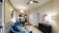 This single-story space showcases a thoughtfully designed open-concept floor plan that connects the living, dining, and kitchen areas into one cohesive, flowing environment. The living area is anchored by dark hardwood-style flooring and features a ceiling fan with a light fixture, contributing to both comfort and ambiance. Neutral-toned walls enhance the sense of openness throughout.

The kitchen is well-appointed with dark cabinetry, stainless steel appliances, and a kitchen island or peninsula that serves as a natural divider between the cooking and dining spaces. Pendant lighting above the island adds a modern, stylish touch, while additional overhead lighting ensures the area is well-lit.

The dining area sits adjacent to the kitchen, creating a practical and social layout ideal for everyday living. A corridor visible from the main living space leads to additional rooms, including what appears to be closet storage and a separate room accessed through a white-paneled door. A dark dresser with decorative accents adds a refined touch near the hallway. Overall, the space presents a modern, well-organized interior with consistent finishes throughout.