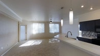 This single-floor residence offers an open-concept layout that connects the kitchen and living area into one cohesive, light-filled space. The living room is finished with plush carpeting and features a ceiling fan, along with large windows dressed with vertical blinds that allow generous natural light to filter through, casting a warm glow across the room. Crown molding adds a refined architectural detail along the upper walls. A private entry door is visible to the side, providing convenient access. The kitchen area is defined by a smooth, light-colored countertop that extends into a peninsula, creating a natural divide between the cooking and living spaces. A stainless steel sink is integrated into the counter, accompanied by a contemporary gooseneck faucet. Overhead, two sleek cylindrical pendant lights hang at varying heights, adding a modern touch and providing focused task lighting. The overall color palette is neutral and bright, with white walls and ceilings enhancing the sense of openness. The space presents a clean, functional design that balances everyday practicality with understated modern style.