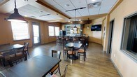 This single-story commercial dining space combines a casual bar and restaurant layout designed for comfortable, flexible use. The main dining area features hardwood-style flooring and a mix of standard and high-top tables paired with wooden ladder-back chairs with cushioned seats, accommodating various group sizes. Warm, tan-toned walls complement the rich wood furnishings, creating an inviting atmosphere.

At the rear of the space, a dark-finished bar serves as the focal point, featuring pendant lighting and a mirrored back bar display illuminated with soft blue lighting, giving it a distinctive visual appeal. A wall-mounted flat-screen television is positioned nearby, making it well-suited for watching sporting events or entertainment.

Overhead, a drop ceiling with recessed and track lighting provides ample, evenly distributed illumination throughout the space. Natural light filters in through windows near the entrance, enhancing the overall brightness. The entry area includes a glass-panel door, offering easy access and visibility to the exterior. The open floor plan allows for straightforward traffic flow between the dining area and the bar, making the space functional and easy to navigate.