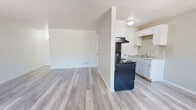 This single-floor unit offers a clean, functional layout with a bright and neutral aesthetic throughout. Light gray wood-look flooring runs continuously through the main living areas, creating a cohesive and modern feel.

The open living area provides generous room for furnishings and benefits from a partially vaulted ceiling that adds a sense of height and airiness. A half-wall partition separates the living space from the kitchen, maintaining visual openness while defining each zone.

The galley-style kitchen is efficiently designed with white upper and lower cabinetry offering ample storage, a countertop with an integrated sink, and black appliances including a range with overhead ventilation and a full-size refrigerator. Natural light filters in, and the white finishes keep the space feeling clean and bright.

The bedroom is a comfortable size, featuring a ceiling fan, a horizontal window that brings in natural light, two closets for generous storage, and built-in open shelving — providing excellent organization options.

The bathroom is finished in crisp white and includes a full soaking tub with an overhead shower, chrome fixtures, and a small window that allows for natural ventilation. The overall design of the unit is clean and move-in ready, with a consistent neutral palette that makes for a welcoming and versatile living space.
