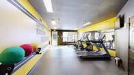 This fitness center is a well-organized, single-room exercise space designed to support a comprehensive workout experience. The interior is defined by a bold two-tone color scheme — energizing yellow accent walls paired with deep charcoal gray surfaces — creating a dynamic and motivating atmosphere. A large motivational mural reading "Don't Make Excuses, Make Results" dominates one wall, reinforcing the space's purpose-driven design.

The exercise room is outfitted with a variety of cardio equipment, including treadmills and elliptical machines, all positioned along a full-length wall mirror that visually expands the space and allows users to monitor their form. On the strength training side, a cable and pulley machine system is paired with a dedicated weight bench and a rack of free weights, providing options for a complete resistance workout. Medicine balls and other functional fitness accessories are also available.

Durable rubber flooring runs throughout, offering both safety and comfort underfoot. Recessed and flush-mounted ceiling fixtures provide bright, even illumination. Natural light filters in through multiple windows, enhancing the open feel of the space. A wall-mounted television adds an entertainment option during workouts. The layout allows for an efficient flow between equipment areas, making the most of the available footprint.