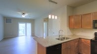 Located at 850 Lisburn Rd in Camp Hill, Pennsylvania, this single-floor residence offers a thoughtfully designed open-concept living space that seamlessly connects the kitchen and living areas. The kitchen serves as the heart of the home, featuring a central island topped with granite countertops, warm honey-toned wood cabinetry, a double basin sink with a modern gooseneck faucet, and a built-in dishwasher. Pendant lighting above the island adds both function and style, illuminating the workspace with a contemporary flair. The open layout flows naturally into the living area, which benefits from hardwood-style flooring that extends throughout. A ceiling fan enhances comfort in the main living space, while vertical blinds frame a sliding glass door that opens to a private balcony, inviting an abundance of natural light and outdoor views. Neutral wall tones and clean architectural lines give the interior a bright, airy ambiance. A hallway visible from the main living area leads to additional rooms, offering a well-organized and functional floor plan suited for everyday living. Overall, the space combines practical design with comfortable, modern finishes.