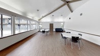 This single-floor community room offers a generous, open layout designed for gatherings and shared activities. The space is defined by its striking vaulted ceiling, which rises dramatically and is accented by exposed wooden beams that add a rustic architectural character to the otherwise clean, white interior. The hardwood-style flooring runs throughout, lending warmth and cohesion to the room.

A row of large windows lines one wall, providing an abundance of natural light and direct views of an outdoor pool area and surrounding courtyard. Additional windows on the opposite wall further brighten the interior, creating an airy and welcoming atmosphere.

The main area is furnished with multiple round white tables and black chairs, arranged to accommodate small groups for meetings, dining, or casual socializing. A dark sofa is positioned along one wall, offering a comfortable seating alternative. A whiteboard is also present, suggesting the space can serve educational or organizational purposes.

A secondary adjoining area is accessible through an interior door and appears to include basic appliances, providing additional utility. The overall ambiance is functional yet comfortable, well-suited for a multi-use community gathering space.