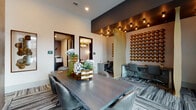 This thoughtfully designed single-floor workspace blends contemporary aesthetics with practical functionality across several distinct zones. The main collaborative area centers on a live-edge wood conference table paired with sleek leather chairs, set atop patterned striped carpeting that runs throughout the space. A striking accent wall adorned with rows of bronze sculptural discs adds warmth and texture, while undulating gold room dividers create visual separation between workstations without fully enclosing them. Individual workstations feature built-in desks beneath bold abstract artwork, offering focused work settings equipped with power access. Private conference rooms are finished with deep navy accent walls and wall-mounted flat-screen displays, ideal for small meetings or presentations. A lounge area with soaring double-height windows floods the space with natural light, offering outdoor views and comfortable seating including tufted blue armchairs and organic stone side tables. Wood-panel wall cladding, exposed Edison-style pendant bulbs, and recessed lighting throughout contribute to an atmosphere that is simultaneously professional and inviting, making this space well-suited for both focused work and collaborative gatherings.