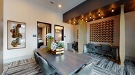 This thoughtfully designed single-floor workspace blends contemporary aesthetics with practical functionality across several distinct zones. The main collaborative area centers on a live-edge wood conference table paired with sleek leather chairs, set atop patterned striped carpeting that runs throughout the space. A striking accent wall adorned with rows of bronze sculptural discs adds warmth and texture, while undulating gold room dividers create visual separation between workstations without fully enclosing them. Individual workstations feature built-in desks beneath bold abstract artwork, offering focused work settings equipped with power access. Private conference rooms are finished with deep navy accent walls and wall-mounted flat-screen displays, ideal for small meetings or presentations. A lounge area with soaring double-height windows floods the space with natural light, offering outdoor views and comfortable seating including tufted blue armchairs and organic stone side tables. Wood-panel wall cladding, exposed Edison-style pendant bulbs, and recessed lighting throughout contribute to an atmosphere that is simultaneously professional and inviting, making this space well-suited for both focused work and collaborative gatherings.