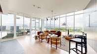 Located at 912 2nd Ave N in Nashville, Tennessee, this sophisticated rooftop amenity space offers a seamless blend of modern interior design and expansive outdoor living. The single-floor living room is bathed in natural light, thanks to soaring floor-to-ceiling glass walls that frame sweeping panoramic views of the Nashville skyline and surrounding landscape, including the Cumberland River.

Inside, wide-plank light hardwood floors anchor a thoughtfully curated lounge area featuring a bold rust-orange sectional sofa, mid-century-inspired accent chairs, and a statement brass pendant light fixture. A striking deep blue shiplap accent wall adds character and warmth, while an integrated wet bar with artisan tile, arched architectural detailing, and open shelving serves as an elegant focal point for entertaining.

The space transitions effortlessly to a generous outdoor terrace, where rows of woven lounge chairs invite relaxation beneath the open sky. Wood-paneled ceiling accents and glass balcony railings define the terrace, which also features a dining area and lush planters. A vibrant large-scale mural punctuates the outdoor space with artistic energy, perfectly complementing the contemporary design aesthetic throughout this premier Nashville rooftop retreat.