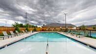 Located at 1800 NW 115th Terr in Oklahoma City, Oklahoma, this outdoor amenity space serves as a central gathering area centered around a generously sized swimming pool with striking blue-tiled edges and crystal-clear water. The pool is accessible via multiple stainless steel entry rails and steps, and is fully enclosed by a black wrought iron safety fence, providing both security and an open sightline across the surrounding landscape.

The expansive concrete pool deck offers ample room for relaxation, lined with rows of lounge chairs positioned along all sides of the pool. Neatly trimmed shrubs, decorative rock beds, and patches of green turf add a polished, landscaped character to the perimeter, softened further by a tall wooden privacy fence that encloses the space.

A standout feature is the attached pool house, constructed with a combination of brick and board-and-batten green siding beneath a gabled roof. This structure provides a covered outdoor patio area equipped with a ceiling fan, outdoor lighting, and direct pool access — ideal for entertaining or seeking shade. Additional outdoor seating and table arrangements are positioned under a separate umbrella area nearby.

The overall design reflects a well-maintained, community-oriented outdoor environment, thoughtfully laid out for both leisure and social use in a residential neighborhood setting.