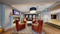 Located at 759 Thornton Way in Alexandria, Virginia, this single-floor amenity space exemplifies upscale communal design with a refined, hotel-inspired aesthetic. The layout flows seamlessly across several distinct zones, each thoughtfully appointed with curated furnishings and cohesive décor.

The entry foyer sets an immediate tone of sophistication, featuring intricate geometric parquet hardwood flooring, a sleek dark credenza with brass hardware, flanking table lamps, and a striking abstract artwork as a focal point. A built-in mailbox wall adds functional elegance nearby.

The primary lounge area centers around cognac leather club chairs arranged around a marble-top coffee table, all anchored by a bold black-and-white geometric area rug. A dramatic navy and gold drum chandelier overhead draws the eye upward, while a classic fireplace with wall-mounted television provides a warm gathering focal point. Wainscoting and crown molding detail the walls throughout.

An adjacent sitting nook features a tufted Chesterfield leather sofa, brass pendant lighting, and large windows inviting ample natural light. A secondary lounge with emerald drapery and street-facing windows adds another intimate retreat.

The game room includes a full billiards table, a stylish wet bar alcove with pendant lighting and vibrant artwork, and glass doors opening directly to an outdoor courtyard, connecting the interior elegantly to the exterior.