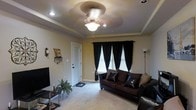 This single-floor space offers a thoughtfully designed interior with standout architectural and decorative details. The living area serves as the centerpiece of the space, showcasing an elegant tray ceiling with recessed lighting and a decorative ceiling fan that adds both style and function. Soft, plush carpeting runs throughout, contributing to a warm and comfortable ambiance. A pair of windows dressed in dramatic black curtains with classic white blinds allows for controlled natural light while adding a bold visual contrast to the neutral, warm-toned walls. The living area is furnished with rich, dark leather seating arranged to create an inviting and functional layout. A sleek media console anchors one wall, while a tall floor lamp provides additional ambient lighting. Decorative accents, including wall-mounted décor and a small potted plant, add personality and a lived-in charm to the space. The interior door and entryway area hint at a well-organized flow throughout the home. Overall, the space combines classic architectural elements with contemporary furnishings to create a cohesive, comfortable, and informative snapshot of single-floor living.