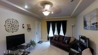 This single-floor space offers a thoughtfully designed interior with standout architectural and decorative details. The living area serves as the centerpiece of the space, showcasing an elegant tray ceiling with recessed lighting and a decorative ceiling fan that adds both style and function. Soft, plush carpeting runs throughout, contributing to a warm and comfortable ambiance. A pair of windows dressed in dramatic black curtains with classic white blinds allows for controlled natural light while adding a bold visual contrast to the neutral, warm-toned walls. The living area is furnished with rich, dark leather seating arranged to create an inviting and functional layout. A sleek media console anchors one wall, while a tall floor lamp provides additional ambient lighting. Decorative accents, including wall-mounted décor and a small potted plant, add personality and a lived-in charm to the space. The interior door and entryway area hint at a well-organized flow throughout the home. Overall, the space combines classic architectural elements with contemporary furnishings to create a cohesive, comfortable, and informative snapshot of single-floor living.