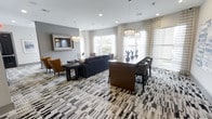 This single-floor space is a thoughtfully designed communal lounge that blends contemporary style with functional comfort. The open-plan layout accommodates both relaxed social gatherings and more structured meetings, anchored by a bold geometric-patterned carpet in neutral tones of black, white, and grey that sets the foundation for the room's sophisticated aesthetic.

A central dining or conference table in rich dark wood is surrounded by upholstered chairs, making it ideal for collaborative work or group dining. Adjacent to this, a plush dark sectional sofa creates a comfortable lounging area, complemented by accent chairs in warm cognac leather that introduce a welcoming contrast to the cooler grey tones of the walls.

Overhead, ornate crystal-style pendant chandeliers are suspended at varying heights, adding a touch of elegance and providing warm, ambient illumination alongside recessed ceiling lights. A wall-mounted flat-screen television is positioned prominently, enhancing the space's versatility as both a social and entertainment hub.

Large windows dressed with sheer striped curtains flood the interior with natural light while maintaining a sense of privacy. The overall ambiance is polished and inviting, combining modern design elements with a warm, accessible atmosphere suited to a variety of uses.