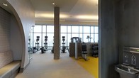 This fitness facility is designed with a clean, modern aesthetic that prioritizes both functionality and an open, energizing atmosphere. The space is anchored by expansive floor-to-ceiling windows that span the length of one wall, flooding the interior with abundant natural light and providing unobstructed views to the outside. The generous ceiling height and open layout contribute to an airy, spacious feel throughout.

Structurally, exposed concrete columns are integrated into the design, adding an industrial character that contrasts effectively with the otherwise polished finishes. The flooring combines light-toned panels with a distinctive yellow rubber section that demarcates the free weights and strength training area, offering both visual clarity and practical safety underfoot.

The cardio zone is well-appointed with a row of upright exercise bikes and treadmills positioned directly in front of the large windows, allowing users to exercise with an open view. Adjacent to this area, a dedicated strength training station features a dumbbell rack and weight bench. Overhead, recessed and panel lighting supplements the natural light, ensuring consistent illumination throughout the facility.