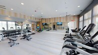 This spacious fitness center is thoughtfully designed to support a comprehensive workout experience. The space features light-toned luxury vinyl flooring throughout, contributing to a clean and modern aesthetic. Walls are finished with a warm, textured stripe wallcovering in neutral tones, complemented by contrasting gray-painted accent walls that add depth and visual interest.

A full-length mirrored wall runs along one side of the room, enhancing the sense of openness while allowing users to monitor their form. Two wall-mounted flat-screen televisions are positioned for easy viewing from multiple workout stations. Recessed lighting and multiple ceiling fans ensure the space remains well-lit and comfortable during use.

The cardio zone features treadmills and elliptical trainers, along with stair-climber machines positioned near oversized windows that offer views of an outdoor pool area and surrounding landscape, providing natural light and a motivating environment. A dedicated functional training area includes a Matrix multi-functional cable machine, a Pilates-style reformer, and an organized rack of medicine balls and resistance accessories. The overall ambiance is bright, airy, and professionally appointed, reflecting a high standard of amenity design.