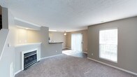 This two-floor space features an open and airy layout, highlighted by a generously sized living area with neutral-toned walls and plush wall-to-wall carpeting that adds warmth underfoot. A classic white fireplace serves as a focal point along one wall, contributing an inviting, cozy atmosphere to the room. Natural light flows in through a large sliding glass door fitted with vertical blinds, which provides access to an outdoor area, as well as through an additional window that further brightens the interior. A ceiling fan is centrally positioned overhead, offering both function and a finishing touch to the living space. The textured ceiling adds subtle architectural detail throughout. Adjacent to the living area, a kitchen counter with a dark laminate surface creates a natural partition, offering a glimpse into the kitchen beyond, where cabinetry is visible. This pass-through design encourages an open connection between the living and kitchen spaces, making the layout well-suited for everyday living and entertaining. The overall ambiance is clean, neutral, and move-in ready, with a practical design that balances comfort and functionality across both floors.