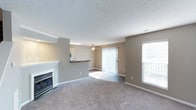 This two-floor space features an open and airy layout, highlighted by a generously sized living area with neutral-toned walls and plush wall-to-wall carpeting that adds warmth underfoot. A classic white fireplace serves as a focal point along one wall, contributing an inviting, cozy atmosphere to the room. Natural light flows in through a large sliding glass door fitted with vertical blinds, which provides access to an outdoor area, as well as through an additional window that further brightens the interior. A ceiling fan is centrally positioned overhead, offering both function and a finishing touch to the living space. The textured ceiling adds subtle architectural detail throughout. Adjacent to the living area, a kitchen counter with a dark laminate surface creates a natural partition, offering a glimpse into the kitchen beyond, where cabinetry is visible. This pass-through design encourages an open connection between the living and kitchen spaces, making the layout well-suited for everyday living and entertaining. The overall ambiance is clean, neutral, and move-in ready, with a practical design that balances comfort and functionality across both floors.