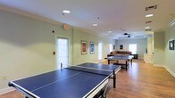 This single-floor recreation space is thoughtfully designed for active leisure and community engagement. The open-plan layout provides generous room for movement and play, accommodating multiple full-sized ping-pong tables arranged side by side across the hardwood floor. Recessed lighting and fluorescent ceiling fixtures ensure the space is well-lit throughout, complemented by abundant natural light streaming in through multiple slatted windows along the walls. A ceiling fan in the far section of the room aids in air circulation, contributing to a comfortable environment during active use. Framed artwork adorns the light green walls, adding a touch of personality and visual interest to the room. Toward the rear of the space, a relaxed lounge area features a comfortable sofa, creating a natural spot for spectators or those looking to unwind. An exit door provides direct access to the exterior, enhancing the room's functionality. Crown molding along the ceiling adds a refined architectural detail that elevates the overall aesthetic. The combination of recreational amenities, casual seating, and bright, open atmosphere makes this a versatile and welcoming communal space.