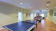 This single-floor recreation space is thoughtfully designed for active leisure and community engagement. The open-plan layout provides generous room for movement and play, accommodating multiple full-sized ping-pong tables arranged side by side across the hardwood floor. Recessed lighting and fluorescent ceiling fixtures ensure the space is well-lit throughout, complemented by abundant natural light streaming in through multiple slatted windows along the walls. A ceiling fan in the far section of the room aids in air circulation, contributing to a comfortable environment during active use. Framed artwork adorns the light green walls, adding a touch of personality and visual interest to the room. Toward the rear of the space, a relaxed lounge area features a comfortable sofa, creating a natural spot for spectators or those looking to unwind. An exit door provides direct access to the exterior, enhancing the room's functionality. Crown molding along the ceiling adds a refined architectural detail that elevates the overall aesthetic. The combination of recreational amenities, casual seating, and bright, open atmosphere makes this a versatile and welcoming communal space.