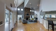 This single-story home makes a striking architectural statement with its soaring vaulted ceilings in the main living area, creating an expansive, airy atmosphere that immediately draws the eye upward. The great room serves as the centerpiece of the home, anchored by a dramatic floor-to-ceiling stacked stone fireplace that serves as both a functional and visual focal point. A flat-screen television is mounted above the fireplace mantel, seamlessly integrating modern technology into the classic design.

Warm hardwood flooring flows throughout the open-concept space, complementing the neutral gray wall tones and white trim work. Recessed lighting and a ceiling fan are installed overhead, while rows of clerestory windows positioned high along the walls allow natural light to filter in from multiple angles. The living area is furnished with plush gray sofas accented with yellow throw pillows, centered over a chevron-patterned area rug.

The layout opens naturally into a dining area toward the rear, where a glass door provides views and access to the outdoors. The overall ambiance is modern yet inviting, blending architectural drama with comfortable, functional design elements.