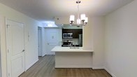 This single-floor residence offers a thoughtfully designed open-concept layout that seamlessly connects the kitchen and living areas. The kitchen is a standout feature, highlighted by dark espresso cabinetry that contrasts beautifully with the crisp white peninsula countertop, which doubles as a breakfast bar and provides additional prep space. Stainless steel appliances — including a refrigerator, electric range, and over-the-range microwave — add a sleek, modern touch. A single-basin sink is integrated into the peninsula, enhancing both functionality and flow. Above, an elegant multi-light chandelier with frosted glass shades serves as a focal point, casting warm, inviting light throughout the space. The flooring throughout is a warm-toned laminate wood, lending a cohesive and contemporary feel to the interior. Bright white walls and recessed lighting contribute to an airy, well-lit atmosphere. A textured ceiling adds subtle character, while clean baseboards and trim throughout speak to the attention to detail in the unit's finishing. The entryway is visible from the main living area, offering a practical and open sense of arrival into the home.