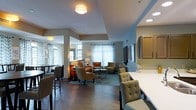 This single-floor space offers an open, flowing layout designed for comfort and social interaction. The interior is anchored by rich, dark hardwood flooring that extends throughout, providing a warm and grounding aesthetic. The main lounge area features a thoughtfully arranged seating arrangement, including upholstered armchairs in neutral and earthy tones, a plush sofa accented with orange throw pillows, and a central coffee table set atop a patterned area rug, creating an inviting conversational zone. A wall-mounted flat-screen television serves as a focal point on one wall, flanked by framed artwork. Large windows dressed with light blue and white chevron-patterned curtains allow generous natural light to fill the room, enhancing its airy ambiance. Recessed lighting and flush-mount ceiling fixtures provide additional illumination throughout. A secondary dining or bar area is visible toward the front, featuring high-top chairs and additional seating beneath pendant-style lighting. Structural columns are integrated seamlessly into the design, subtly defining zones while preserving the open feel of the space. The overall atmosphere is modern yet welcoming, with a carefully curated color palette of grays, blues, and warm neutrals that lends a sophisticated, contemporary character to the environment.