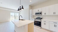 This single-floor unit showcases a thoughtfully designed layout with modern finishes throughout. The kitchen serves as a central highlight, featuring white shaker-style cabinetry with matte black hardware, light quartz countertops, and a full suite of stainless steel appliances including a gas range, over-the-range microwave, and refrigerator. A central island with an integrated sink provides additional prep space and is accented by a striking multi-pendant black light fixture. Light hardwood-style flooring runs throughout the main living areas, complementing the bright, neutral color palette on the walls and ceilings.

The living area opens directly to a private balcony through sliding glass doors, offering outdoor views of surrounding greenery. Natural light flows generously through large windows and the glass door, enhancing the open and airy atmosphere of the space.

The bedrooms feature plush carpeting and sizable windows that bring in additional natural light while framing views of the exterior landscape. The bathroom is finished with a white vanity, quartz countertop, matte black fixtures, a large mirror, and a tub-shower combination, maintaining the cohesive, contemporary aesthetic found throughout the unit.