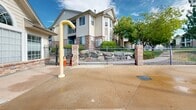 Located at 10500 Irma Dr in Northglenn, Colorado, this outdoor community amenity space is thoughtfully designed to serve as a central gathering hub. The centerpiece is a generously sized freeform swimming pool with a smooth, curved shape that invites leisurely swimming and relaxation. Directly adjacent to the pool sits an attached spa, complete with a decorative water fountain feature that adds a pleasant visual and auditory element to the environment.

The surrounding sundeck is finished in textured aggregate concrete, providing a slip-resistant and durable surface ideal for outdoor use. Ample lounge seating is arranged throughout the deck, with multiple chairs grouped beneath large market umbrellas, offering shaded areas for comfort on sunny Colorado days. A splash pad or water play feature is also present, making the space family-friendly.

The entire area is enclosed by a well-maintained wrought iron fence with brick pillar accents, creating a defined and secure boundary. A gated entry point leads into the pool courtyard, with decorative rock landscaping flanking the entrance. Mature trees and green landscaping are visible throughout the surrounding grounds, providing natural shade and a pleasant aesthetic backdrop. A clubhouse or amenity building with architectural columns and large windows is situated at one end of the complex, completing this well-appointed outdoor retreat.