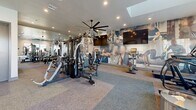 Located at 13215 W Glendale Ave in Glendale, Arizona, this fitness center offers a thoughtfully designed exercise space across a single floor. The room is finished with neutral-toned carpet tiles that define distinct workout zones, while bright recessed lighting and a large ceiling fan ensure a comfortable environment during exercise. Clean white walls are punctuated by bold, large-scale athletic murals that energize the space and reinforce a motivating atmosphere.

The facility is comprehensively equipped to support a full range of workout routines. A multi-station cable and strength machine serves as a centerpiece for resistance training, complemented by a dedicated dumbbell rack and adjustable weight benches. Cardio options include treadmills, elliptical trainers, and stationary bikes. Multiple wall-mounted flat-screen televisions are positioned throughout for entertainment during workouts.

Full-length mirrors along one wall enhance the sense of openness while providing users with proper form feedback. A built-in dual water fountain station offers added convenience. Natural light enters through windows along one wall, and glass doors connect the space to an adjacent area, maintaining an open and airy feel. The overall design strikes a balance between functionality and modern aesthetic, making it a practical and inviting fitness destination.