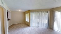 This single-floor residential space offers a comfortable and functional layout designed to maximize natural light and livability. The main living area is a standout feature, highlighted by expansive sliding glass doors dressed with full-length vertical blinds that allow for flexible light control while providing access to the outdoors. A secondary window further contributes to the bright, airy atmosphere of the room. The flooring throughout is soft, wall-to-wall carpeting in a neutral tone, lending warmth and a cohesive feel to the space. Walls are finished in a warm beige palette that complements the light-colored carpet, creating a welcoming and understated ambiance. The textured ceiling adds a classic residential touch, and a flush-mount ceiling light fixture provides practical illumination. Clean baseboards and simple, unadorned walls give the space a blank-canvas quality, making it easy to personalize. Overall, this space combines practicality with an open, light-filled character that suits a variety of living arrangements.