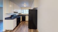 Located at 1539 Tavern Road in Alpine, California, Unit 87 is a single-floor residence that offers a practical and comfortable layout. The kitchen is well-appointed with white cabinetry, dark granite-style countertops, and a full suite of black appliances including a range, dishwasher, and refrigerator. A window above the sink brings in natural light and offers views of surrounding greenery, creating a bright and functional workspace. The main living area features wood-look plank flooring, a textured ceiling, and a large window with blinds that allows ample natural light to fill the room. A wall-unit air conditioner provides climate control, and an open pass-through to the kitchen creates a connected, open feel. A carpeted room with a sliding glass door leads to an outdoor area, flooding the space with sunlight through vertical blinds. Additional rooms include a carpeted bedroom and a secondary room with wood-look flooring, both offering versatile living potential. The bathroom is finished with a white vanity, dark countertop, a large mirror, wood-look flooring, and a full tub-shower combination. Throughout the unit, a neutral color palette and clean finishes provide a move-in ready environment suited to a variety of lifestyles.