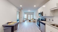 This contemporary single-floor residence offers a thoughtfully designed open-concept layout that seamlessly connects the kitchen, dining, and living areas. The kitchen is outfitted with crisp white shaker cabinetry, granite countertops, a white subway tile backsplash, and a full suite of stainless steel appliances including a range, microwave, dishwasher, and refrigerator. Light wood-style flooring flows throughout the main living space, enhancing the bright, clean aesthetic created by recessed lighting and white walls.

The living area is generous and inviting, anchored by French doors that open to a private balcony, allowing natural light to fill the room and providing outdoor views. A side window further contributes to the airy atmosphere.

The bedroom is a comfortable retreat finished with plush carpeting, a window with exterior views, and ample space for furnishings. The bathroom features a wide granite-topped vanity with white cabinetry, a large mirror, and light wood-style flooring that mirrors the rest of the unit, creating a cohesive design throughout. A closet with paneled doors offers convenient storage. Overall, the space presents a modern, neutral palette with clean lines and quality finishes throughout.