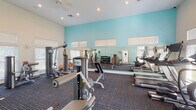 This fitness center in Lafayette, Louisiana, offers a comprehensive workout environment designed to support a wide range of exercise routines. The space is housed on a single floor and is characterized by soaring vaulted ceilings that contribute to an open, airy atmosphere. Walls painted in a fresh aqua-teal tone are complemented by crisp white trim, creating a bright and energizing environment.

Natural light filters in through multiple windows fitted with horizontal blinds, further enhancing the welcoming ambiance. A wall of floor-to-ceiling mirrors spans one side of the room, visually expanding the space and aiding exercisers in monitoring their form. The flooring throughout consists of durable, patterned commercial carpet.

The cardio section is well-appointed with a row of treadmills from True and Matrix brands, along with elliptical trainers and a stationary bike, each equipped with individual TV monitors. The strength-training area includes a selection of cable and resistance machines suited for both upper and lower body workouts, as well as a flat utility bench. A dedicated free-weight station features a multi-tiered dumbbell rack stocked with a variety of weights. Additional amenities include dual water fountains mounted along one wall and a sanitizing wipe dispenser, ensuring a clean and convenient workout experience.