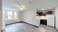 This efficiently designed studio unit offers a single-floor layout that makes excellent use of its intimate footprint. The open main living area features durable light-toned hardwood-style flooring, crisp white walls, and a ceiling fan with integrated lighting that ensures comfortable airflow and illumination throughout. A baseboard heating unit runs along the exterior wall, and a window allows natural light to fill the space.

The kitchen is positioned in an open-plan arrangement adjacent to the living area, featuring rich dark-stained cabinetry that provides generous upper and lower storage. White appliances — including a refrigerator, electric range, and sink — are neatly integrated into the layout, complemented by light countertops and under-cabinet lighting.

The bathroom is finished with full white ceramic tile surrounding a shower-tub combination. Stainless steel grab bars are installed along the tub walls, adding a practical and accessible design element. A frosted window provides ventilation and natural light while maintaining privacy.

A dedicated closet offers a thoughtful organizational system, complete with wire shelving units and a hanging rod, maximizing storage capacity within the space. The unit's straightforward, functional design provides a clean and livable environment well-suited for efficient single-occupant living.
