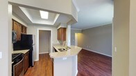 This single-floor residence offers a thoughtfully designed open-concept layout that seamlessly connects the kitchen and living areas, creating a spacious and inviting atmosphere. The kitchen serves as the heart of the home, anchored by a curved peninsula with light granite countertops that provide generous prep and seating space. Warm-toned cabinetry adds a classic touch, while the stainless steel sink and black refrigerator offer functional contrast. A striking skylight positioned above the kitchen floods the space with natural light, enhancing the airy feel of the vaulted ceiling detail overhead. Rich hardwood flooring flows throughout, tying the open living and kitchen zones together with warmth and cohesion. Crown molding trims the ceilings throughout, lending an elegant finishing detail to each room. Neutral gray walls provide a versatile backdrop suited to a variety of décor styles. A doorway off the kitchen leads to a utility or storage area, adding everyday practicality to the layout. The living area extends beyond the kitchen, offering a generous, light-filled retreat. Overall, the space balances comfort and style through its clean lines, quality finishes, and well-considered design.