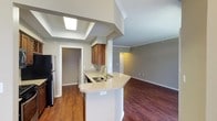 This single-floor residence offers a thoughtfully designed open-concept layout that seamlessly connects the kitchen and living areas, creating a spacious and inviting atmosphere. The kitchen serves as the heart of the home, anchored by a curved peninsula with light granite countertops that provide generous prep and seating space. Warm-toned cabinetry adds a classic touch, while the stainless steel sink and black refrigerator offer functional contrast. A striking skylight positioned above the kitchen floods the space with natural light, enhancing the airy feel of the vaulted ceiling detail overhead. Rich hardwood flooring flows throughout, tying the open living and kitchen zones together with warmth and cohesion. Crown molding trims the ceilings throughout, lending an elegant finishing detail to each room. Neutral gray walls provide a versatile backdrop suited to a variety of décor styles. A doorway off the kitchen leads to a utility or storage area, adding everyday practicality to the layout. The living area extends beyond the kitchen, offering a generous, light-filled retreat. Overall, the space balances comfort and style through its clean lines, quality finishes, and well-considered design.