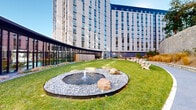 Located at 5159 Pooks Hill Road in Bethesda, Maryland, this thoughtfully designed outdoor courtyard offers a dynamic and well-appointed communal space set between contemporary multi-story buildings clad in light brick and floor-to-ceiling glazing. The grounds are organized around a series of curving pathways that guide visitors through distinct zones, creating a sense of discovery and flow throughout the space.

Lush landscaped planting beds feature ornamental grasses, native shrubs, young deciduous trees, and seasonal perennials that add texture and natural color throughout the year. Stone retaining walls rise along the perimeter, backed by mature trees that provide a natural wooded backdrop and a sense of seclusion despite the urban setting.

A dedicated children's playground area is equipped with natural wood climbing structures, rope ladders, and safety surfacing, offering an engaging play environment. Multiple outdoor seating areas with contemporary tables and chairs are arranged on paver patios, ideal for casual gathering or relaxation. Additional benches are positioned along the walking paths.

Building entrances open directly onto the courtyard through large glass doors framed in dark brick, creating a seamless indoor-outdoor connection. Sleek light poles and ground-level lighting illuminate the space after dark. The overall design balances modern architectural elements with natural landscaping to foster a welcoming, functional outdoor environment.