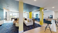 This generously sized single-floor amenity space offers a dynamic and thoughtfully designed environment across a wide-open, interconnected layout. The interior is characterized by a bold contemporary aesthetic, featuring striking yellow accent columns, teal-painted slatted ceilings, and exposed industrial ductwork that add visual interest and a modern edge throughout.

The central lounge area anchors the space with plush sectional sofas arranged on deep blue area rugs over light wood-style flooring, creating a comfortable and inviting gathering zone. Adjacent to the lounge, a bar-height dining counter with vibrant blue stools and pendant lighting offers a casual dining and social setting.

A dedicated gaming area includes a shuffleboard table and a poker/card table set against a bold blue accent wall adorned with decorative wall art, with floor-to-ceiling windows providing abundant natural light and outdoor views of a pool area and surrounding neighborhood.

A co-working zone features built-in desk stations, a communal high-top table with seating, and glass-walled rooms for focused or collaborative work. The entry corridor showcases an architectural open shelving unit with integrated greenery and a media display, serving as a striking focal point. Throughout, large windows and glass doors connect the interior to outdoor balcony access, ensuring a bright and airy ambiance.
