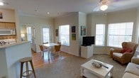 This single-floor residence offers a thoughtfully designed open-concept layout that seamlessly connects the living and dining areas into one cohesive, comfortable space. Large windows throughout flood the interior with abundant natural light, creating an airy and inviting atmosphere. The dining area features hardwood flooring and accommodates a modest dining table with seating for four, positioned conveniently near a set of glass-paneled exterior doors that provide additional light and outdoor access. The living area transitions to carpeted flooring and is centered around a flat-screen television, with ample space for comfortable furnishings. A ceiling fan with a light fixture is installed overhead, providing both lighting and climate comfort. The walls are finished in a clean, neutral tone that complements the natural light and gives the space a fresh, open feel. Crown molding adds a subtle architectural detail throughout. Colorful artwork displayed on the walls adds character and visual interest to the otherwise understated interior. The overall ambiance is clean, functional, and welcoming, making it a practical and pleasant everyday living environment.