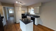 This two-floor residence offers a thoughtfully designed layout with functional living spaces across both levels. The kitchen stands out as a central highlight, featuring crisp white cabinetry that extends to the ceiling for generous storage capacity. Dark granite countertops provide a striking contrast against the white cabinets, running along the perimeter and continuing onto a freestanding center island — ideal for additional prep space or casual dining. Black appliances, including a range and dishwasher, complement the kitchen's bold color palette. A stainless steel sink is positioned beneath a window, allowing natural light to filter into the space. Track lighting overhead ensures the kitchen remains well-lit throughout the day. Warm hardwood flooring flows throughout, adding richness and continuity to the overall aesthetic. A glimpse through the adjoining doorway hints at the additional living areas connected to the kitchen, suggesting an open and accessible floor plan. The neutral wall tones throughout create a clean, versatile backdrop that suits a variety of interior styles. With its combination of classic design elements and practical features, this home offers a comfortable and inviting environment across both of its floors.
