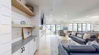 Located at 16 Congress St in Beverly, MA, this open-concept kitchen and living space offers a thoughtfully designed single-floor layout that blends modern coastal style with warm architectural details. Striking exposed wood beam ceilings with pendant lighting anchor the kitchen area, where crisp white shaker cabinetry, a quartz-topped island with seating for multiple guests, and a eye-catching blue and white patterned tile backsplash create a vibrant focal point. Open metal and wood shelving adds both function and character, while a built-in beverage cooler and undermount sink complete the well-appointed kitchen. The living area features a sleek gas fireplace set within a shiplap-clad accent wall, flanked by built-in shelving and cabinetry. Light hardwood flooring flows throughout, enhancing the sense of openness and warmth. A dedicated lounge zone with large floor-to-ceiling sliding glass doors and expansive windows floods the space with natural light and provides access to an outdoor area with exterior views. A pool table rounds out the entertainment offerings, making this a versatile and inviting communal space suited for relaxation and social gatherings alike.