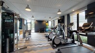 This fitness center is a thoughtfully designed single-level space that balances functionality with contemporary aesthetics. The room is anchored by a striking chevron-patterned carpet in neutral tones of gray, gold, and beige, lending visual energy to the floor throughout the entire space.

Large floor-to-ceiling windows line multiple walls, flooding the interior with natural light and offering outdoor views that enhance the workout environment. The ceiling features a clean white paneled finish with pendant-style industrial lights suspended at regular intervals, complementing the recessed lighting for a well-lit atmosphere. Deep navy and teal accent walls provide a bold, modern contrast to the bright, airy feel of the space.

The cardio zone is well-stocked with treadmills, elliptical machines, and a stationary bike, all positioned to face the expansive windows. A dedicated strength training area includes a multi-station cable machine, free weights on a rack, and additional resistance equipment. A stretching and functional fitness corner features exercise balls and open floor space, while a full-length mirror along one wall visually expands the room. A wall-mounted television and a built-in water fountain station with a sanitizing dispenser add practical amenities to the overall experience.