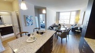 This single-floor residence showcases a thoughtfully designed open-concept layout that seamlessly connects the kitchen, dining, and living areas. The kitchen features a generously sized granite-topped island with dark cabinetry and brushed nickel hardware, providing ample prep space and casual seating. Pendant lighting hangs above the island, adding a warm, focused glow to the space.

The dining area is positioned centrally, anchored by a round glass-top table surrounded by upholstered chairs, creating an elegant yet functional gathering spot. Large windows along the far wall flood the living area with natural light, enhancing the airy, open feel of the space. Warm hardwood-style flooring runs throughout, providing a cohesive visual flow between zones.

A neutral color palette of soft grays and whites keeps the interior feeling bright and contemporary. Colorful geometric wall art adds a subtle decorative accent near the kitchen. A glimpse through an open doorway reveals a bedroom beyond, suggesting a well-connected and efficiently organized floor plan. Overall, the space balances modern finishes with a comfortable, livable ambiance.